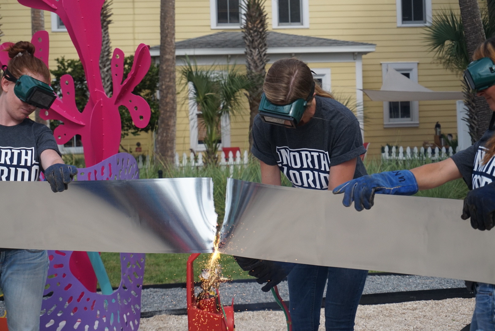 In lieu of a ribbon, UNF sculpture student Gillian Harper blazes through a strip of metal with a blowtorch with the help of Emily Pinnell (left) and Mary Ratcliff (right). Not pictured: Diana Shepherd (far left) and David Peters (far right).